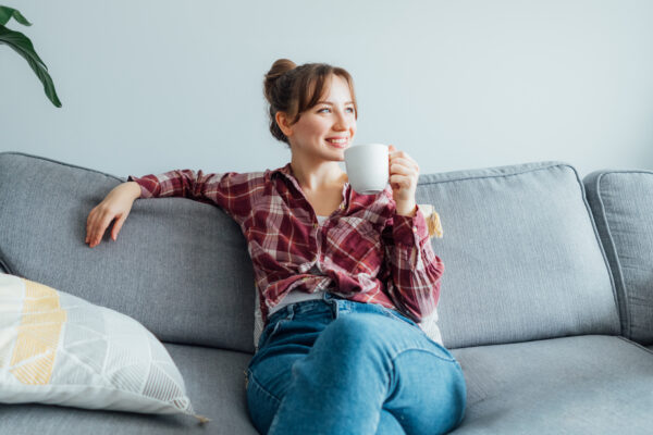 A girl sitting on her couch with a cup of coffee, looking off into the distance, smiling.