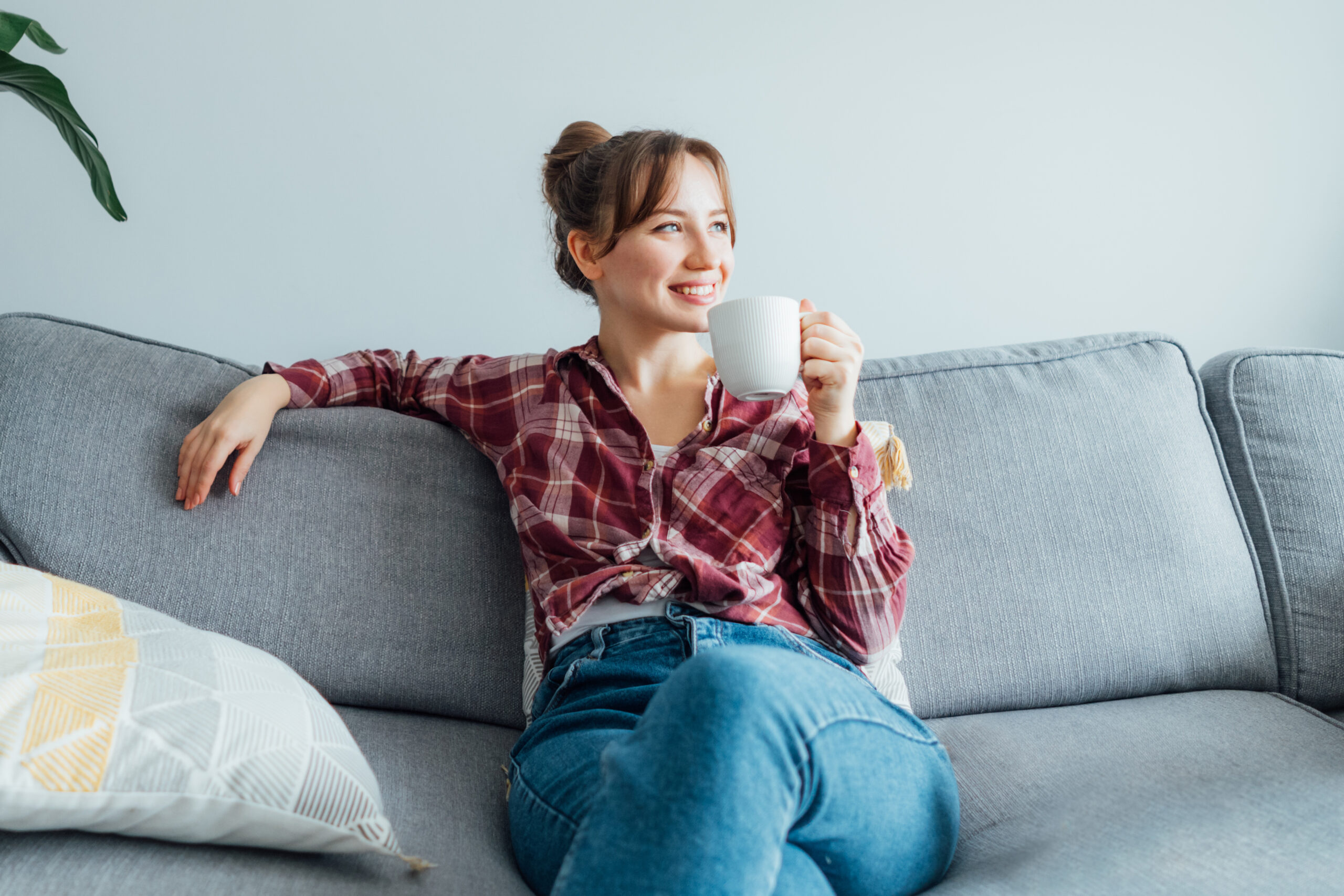 A girl sitting on her couch with a cup of coffee, looking off into the distance, smiling.