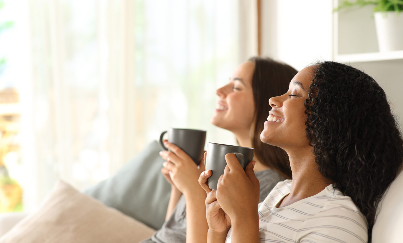 Two young women sitting side by side, enjoying the fresh air in the home, while holding coffee mugs.