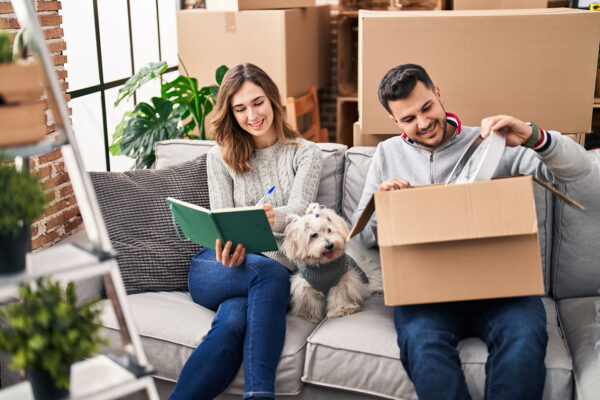 A couple and their dog sitting on a couch, unpacking a cardboard box with a clock and writing in a notebook about what they are working on.