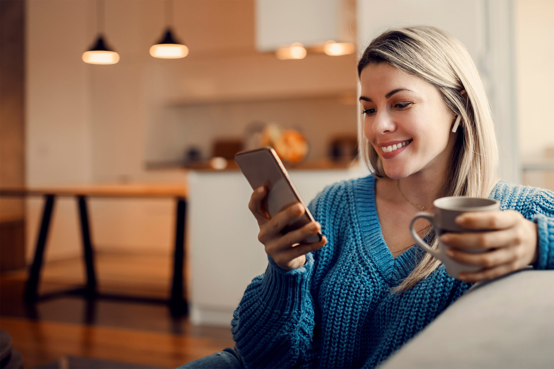 A happy woman hanging on social media on the phone and drinking coffee at home.